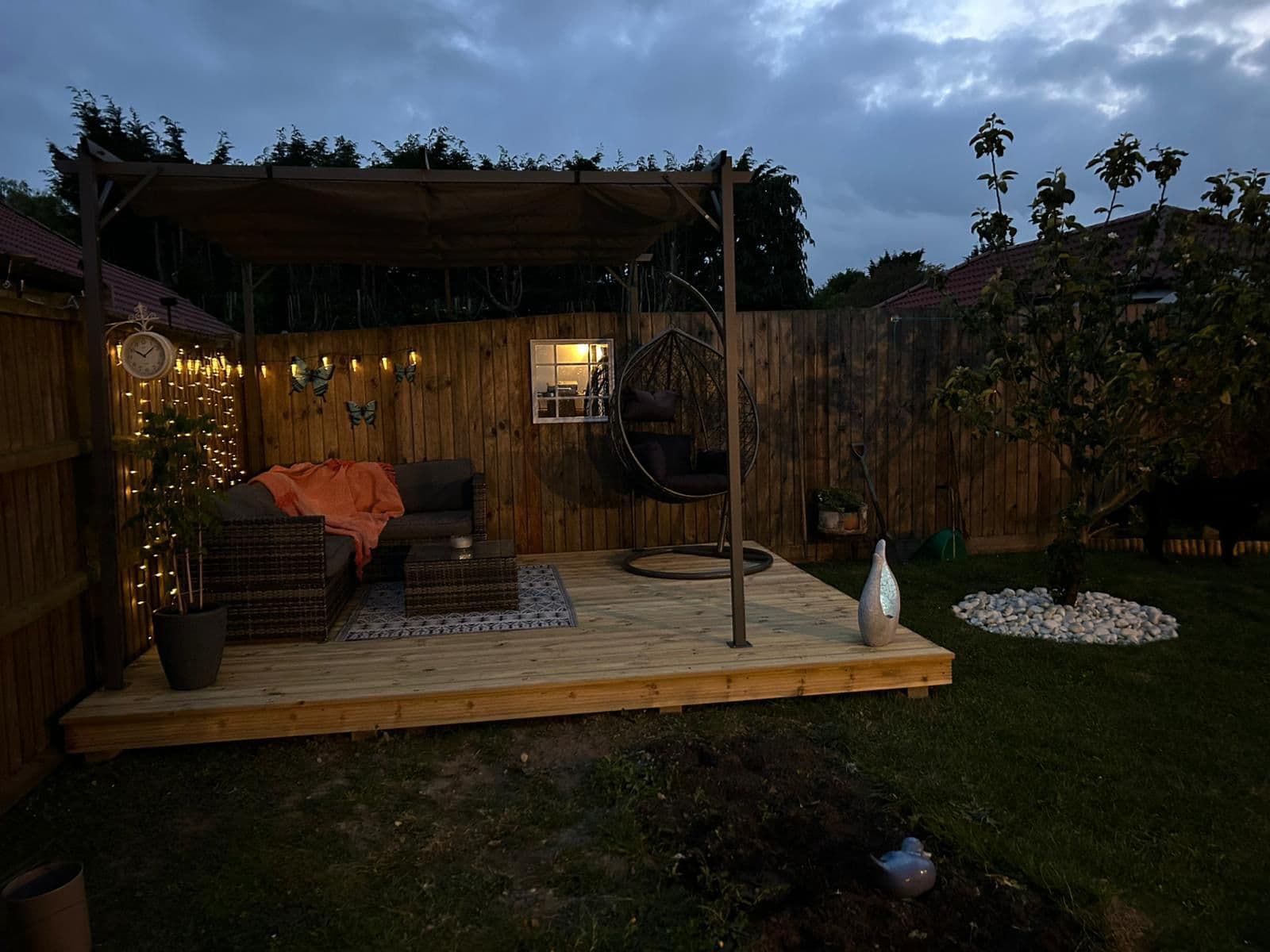 Cozy wooden deck at dusk with a wicker sofa, hanging chair, and warm string lights.